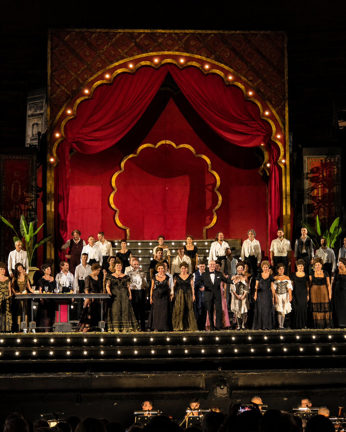 The full company beneath the production’s red cabaret arch — Paul Curran’s Moulin Rouge-inspired Paris turning Violetta’s world into public spectacle before the tragedy closes in.