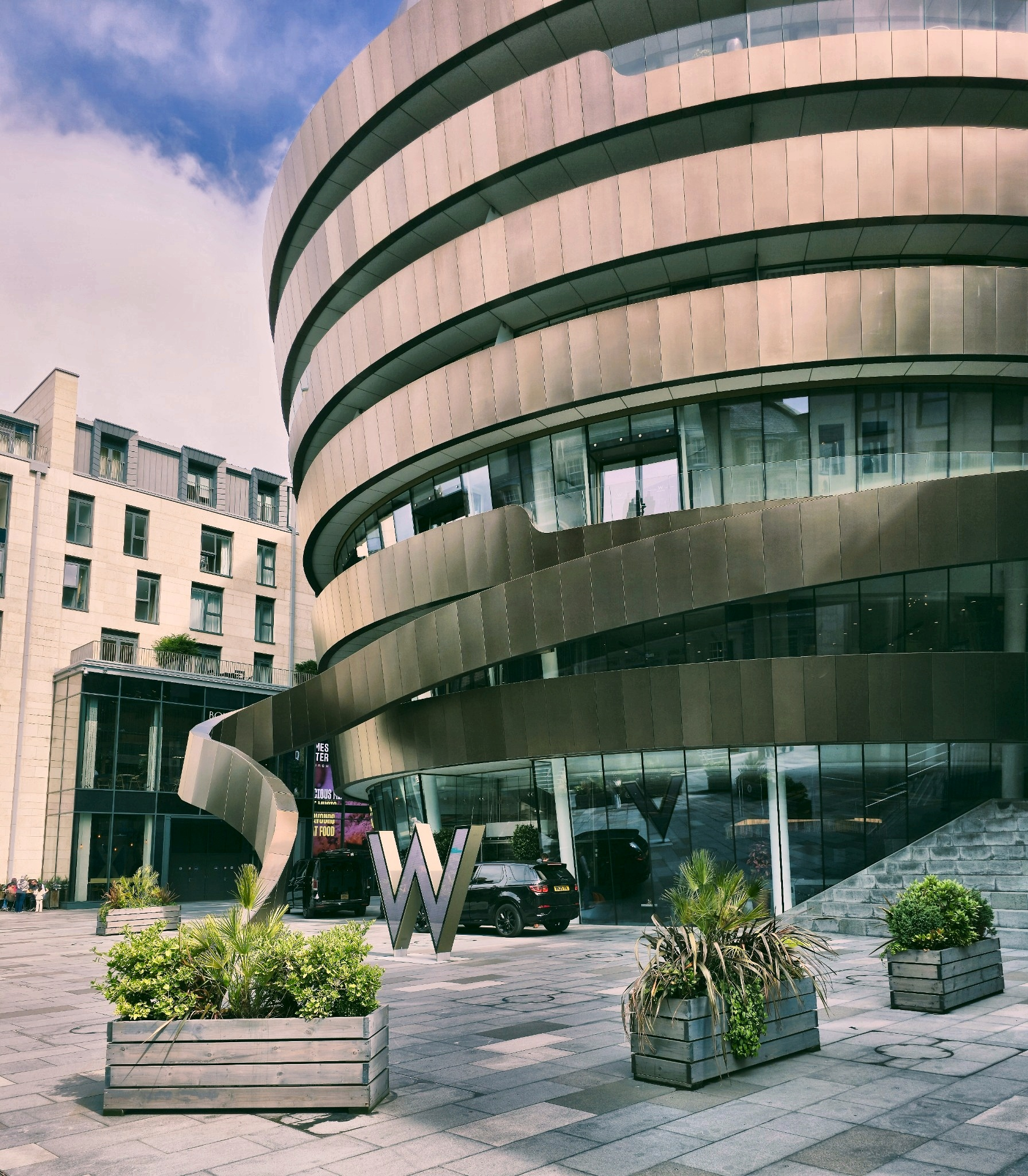 By daylight, W Edinburgh’s bronze ribbon wraps itself around St James Quarter with considerably more confidence than restraint.