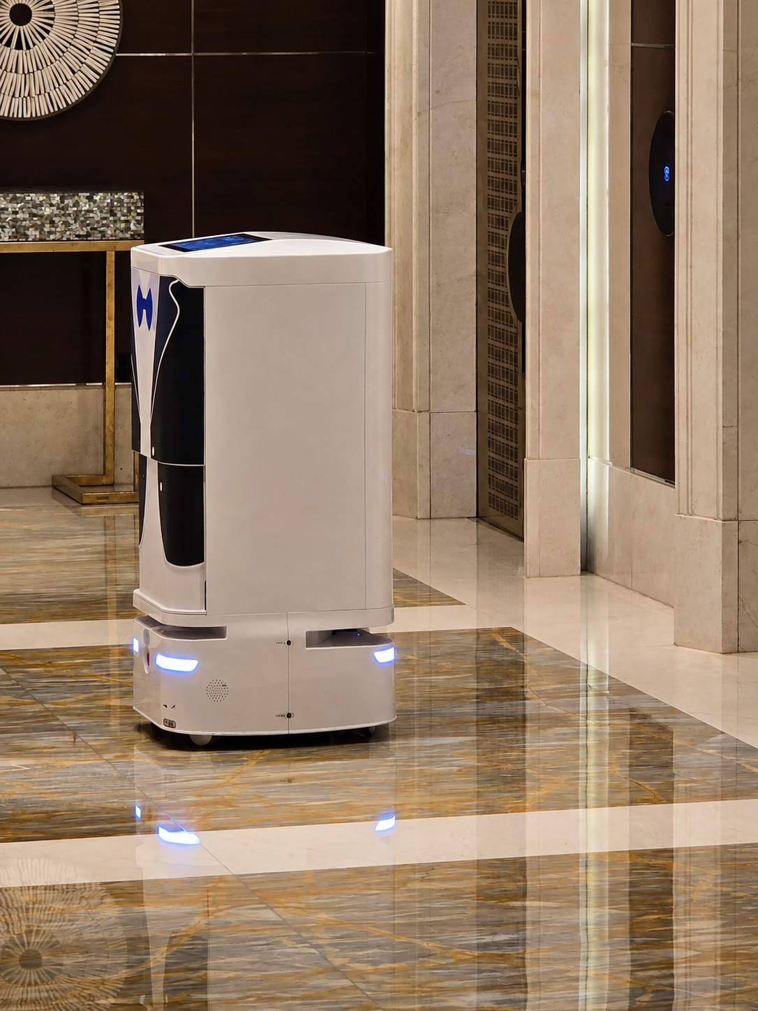 A white room-service robot with blue lights waits beside the lifts in a hotel lobby.