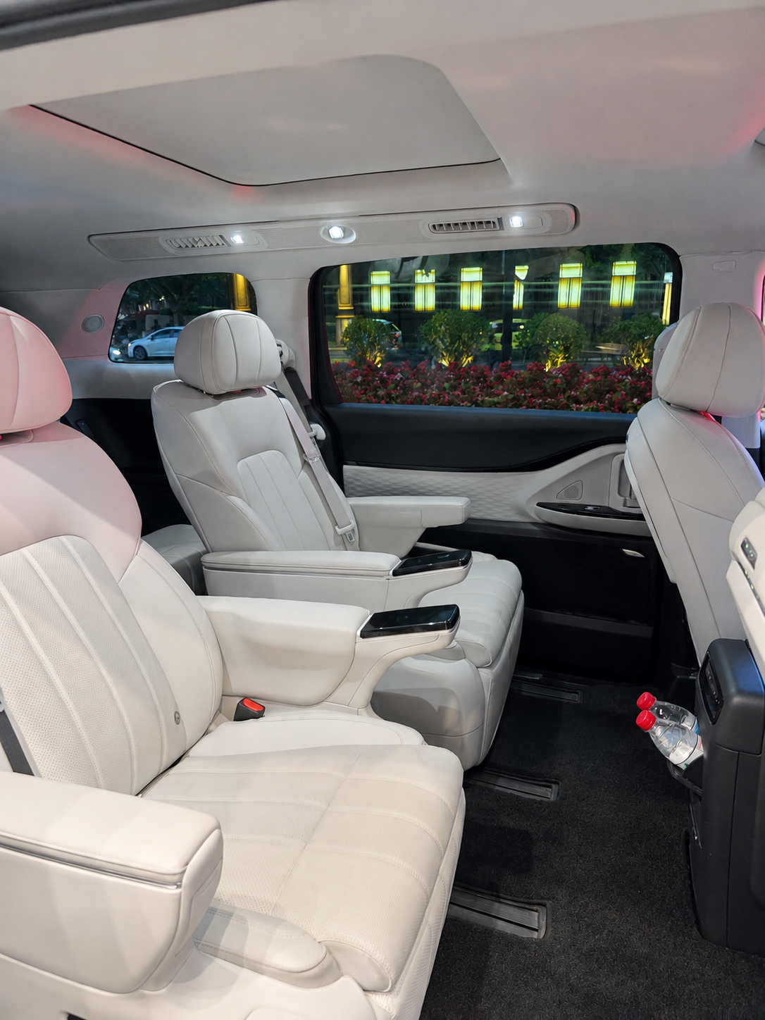 White leather passenger seats inside a luxury vehicle, with a flower bed and illuminated building visible outside.