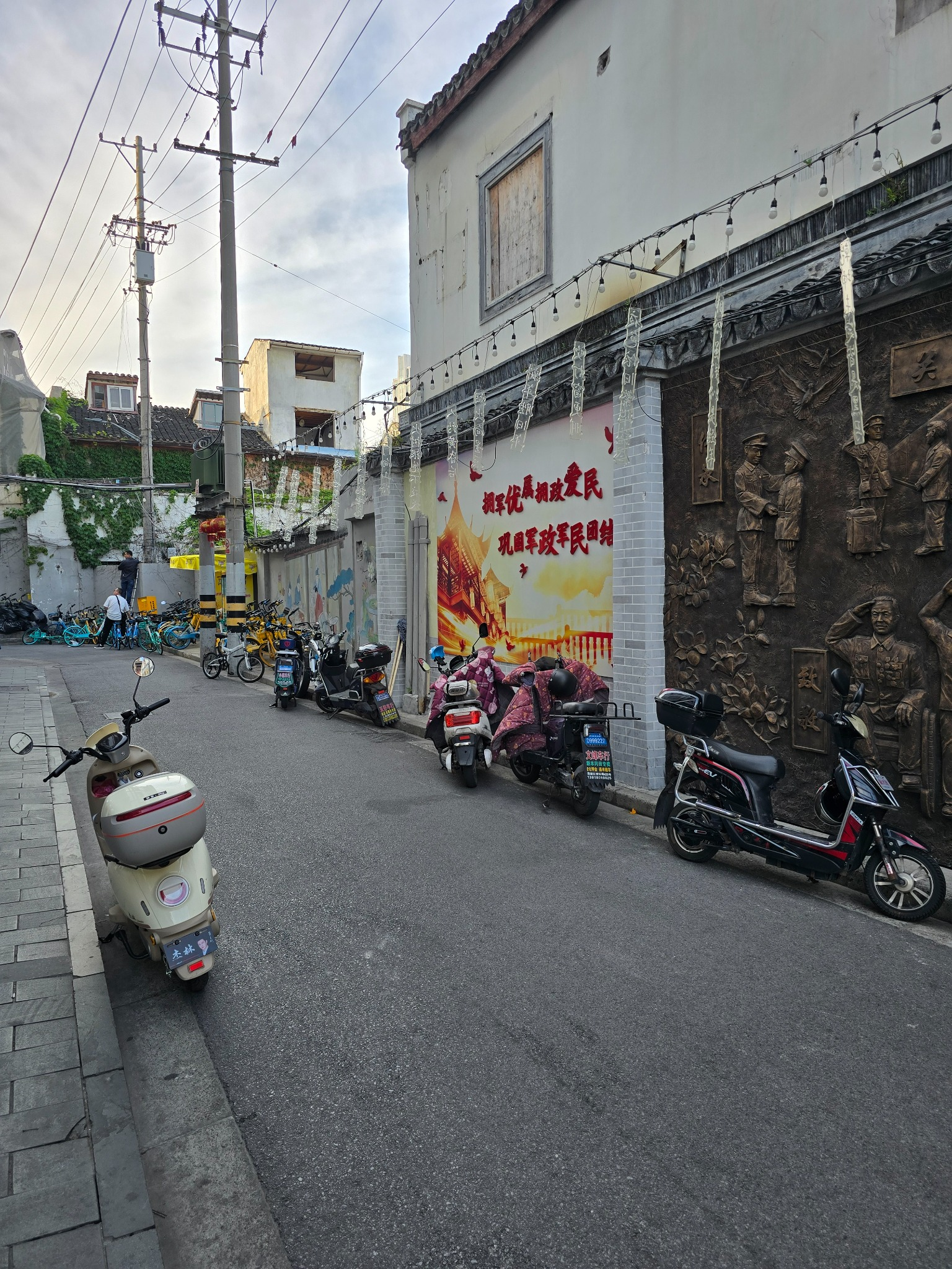 Parked scooters line a narrow Shanghai street beside a traditional building with a large Chinese mural.