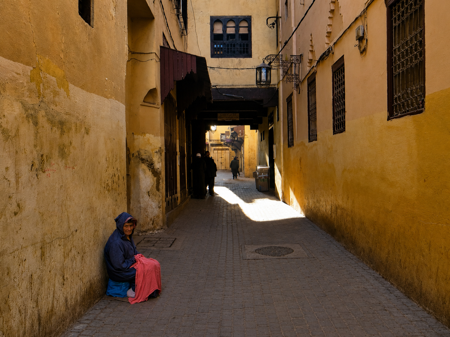 Seated person in a blue top and pink skirt beside a narrow, sunlit medina alley in Meknes.