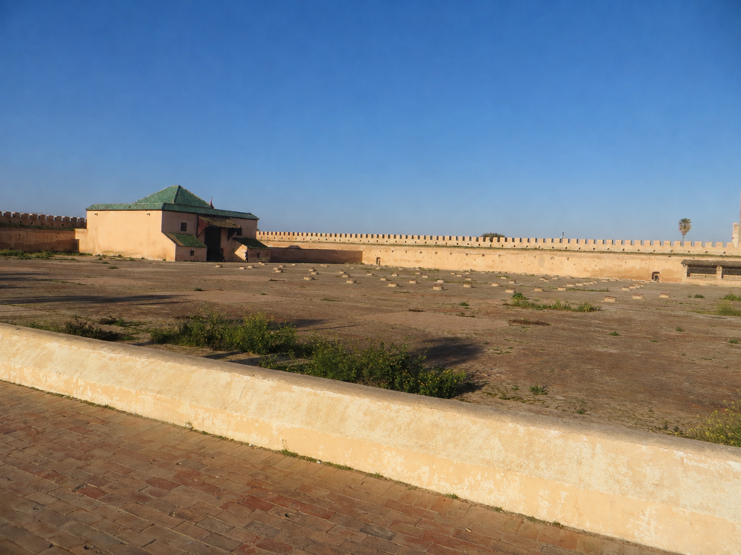 Crenellated wall and green-roofed gate border an open courtyard in Meknes.