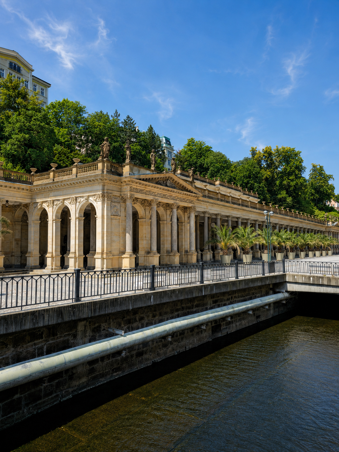 The Mill Colonnade lines the Teplá River in Karlovy Vary beneath a blue sky.