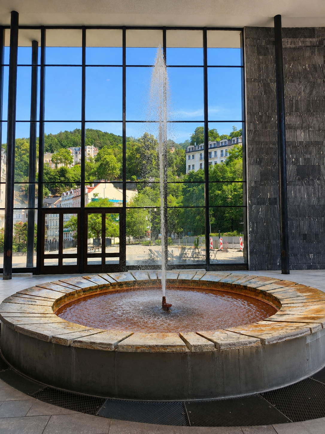 Vřídlo hot spring fountain inside a modern hall in Karlovy Vary, with hills and buildings visible beyond.