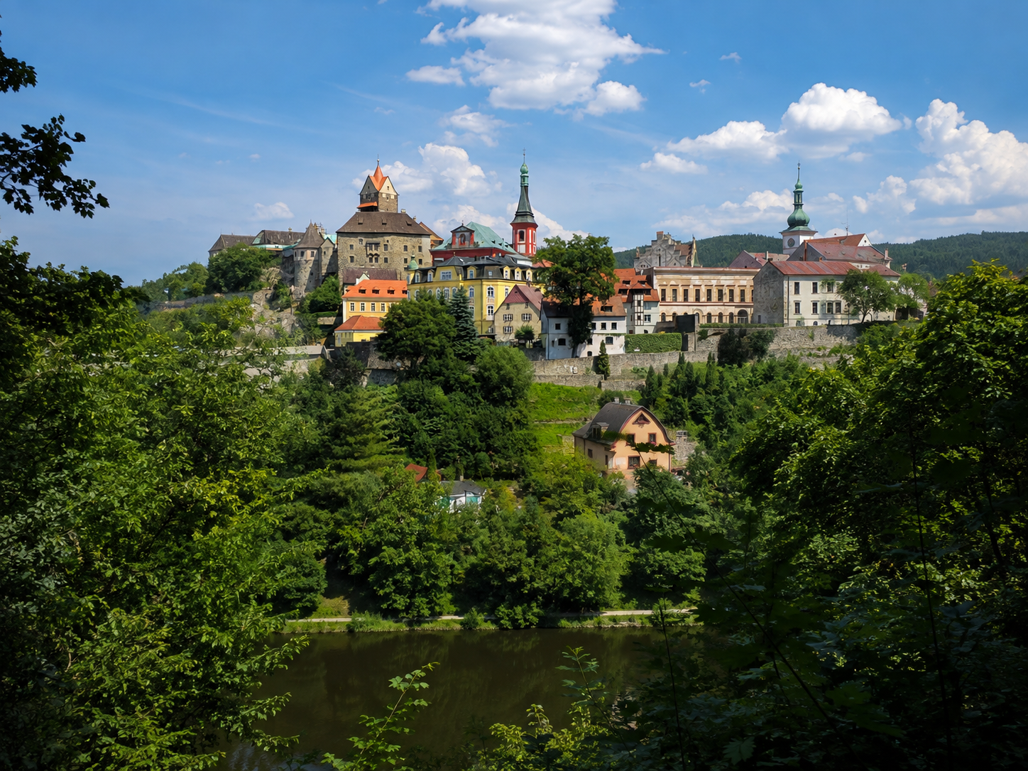 Loket Castle and church towers above the Ohře River, viewed across wooded banks.