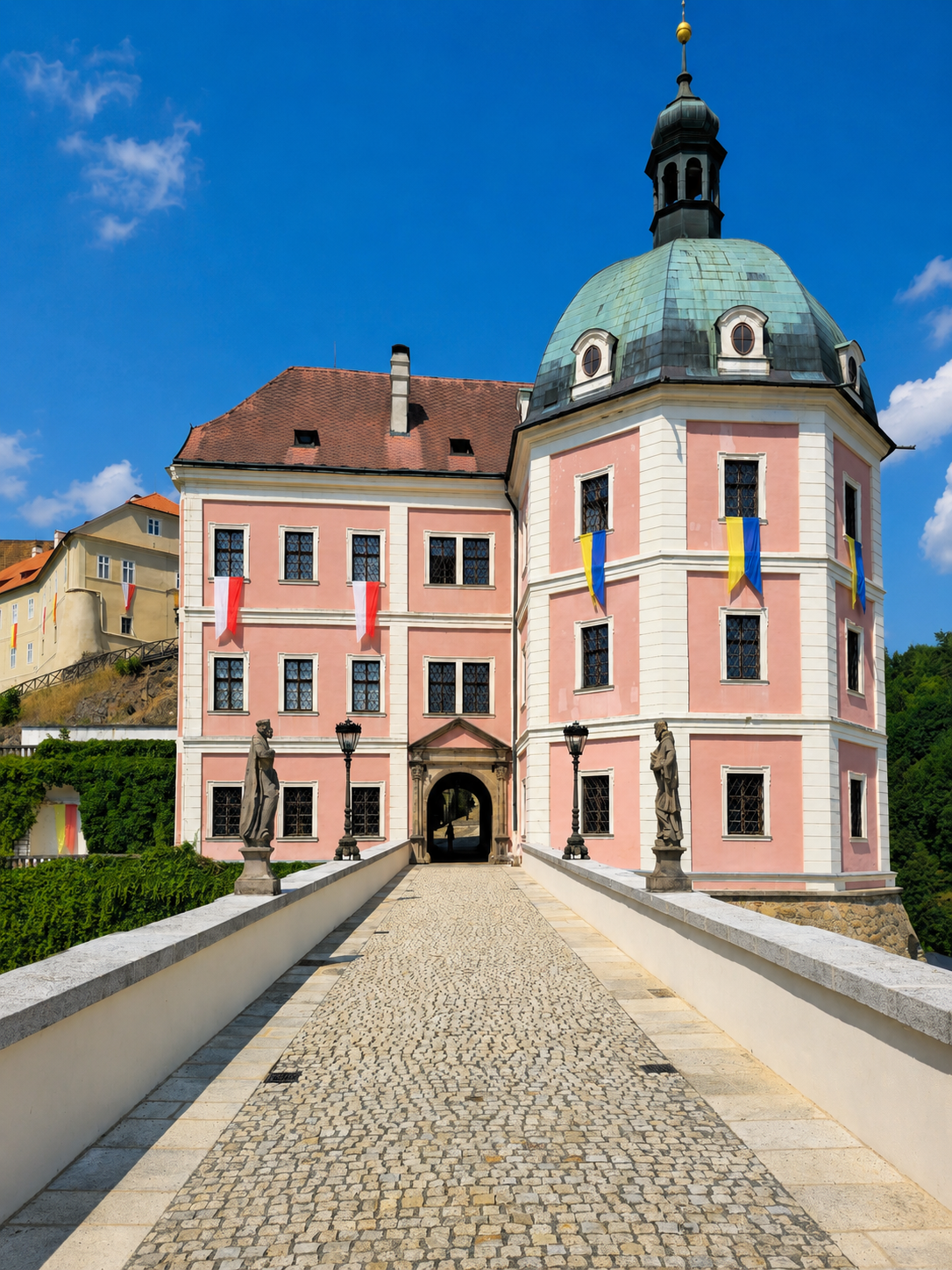 Pink Baroque chateau with a domed tower and flags, viewed from a cobblestone bridge in Bečov.