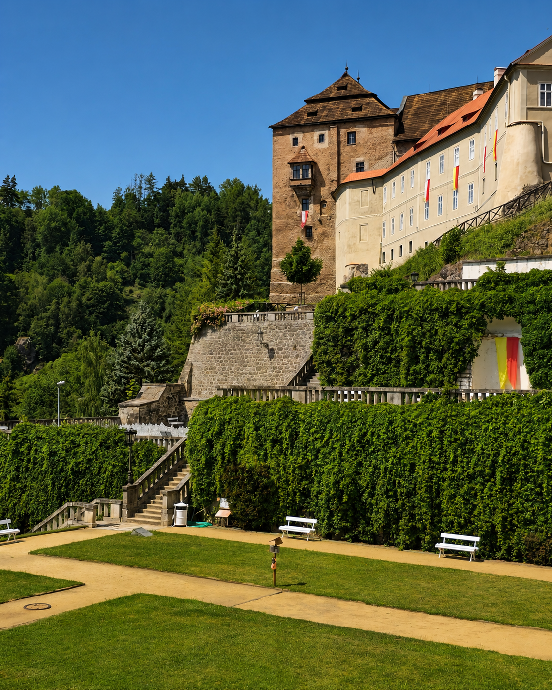 Bečov Castle above formal gardens, with ivy-covered walls, paths, benches, and a wooded hillside.