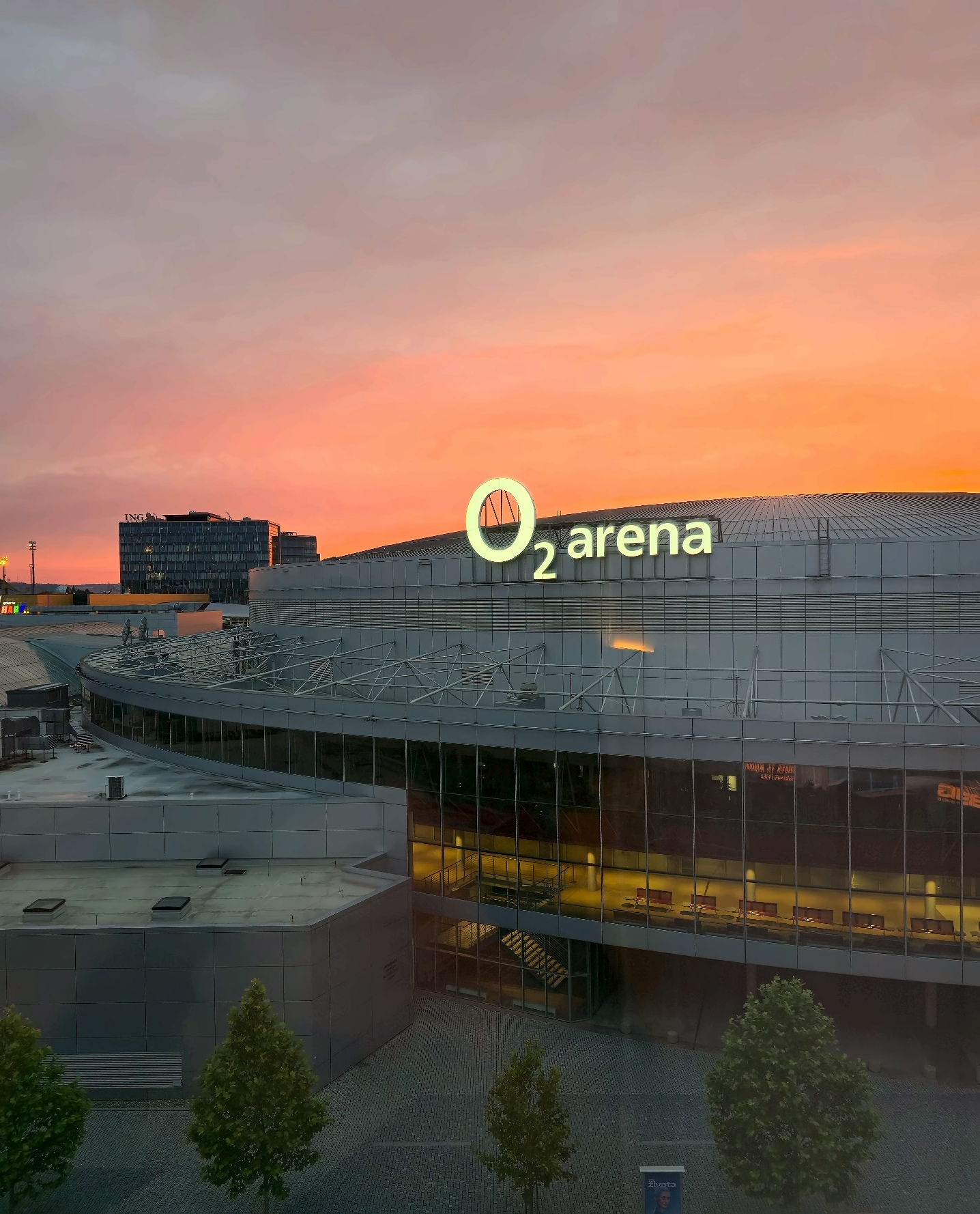 O2 Arena with its illuminated sign and curved roof under a pink-orange sunset sky in Prague.