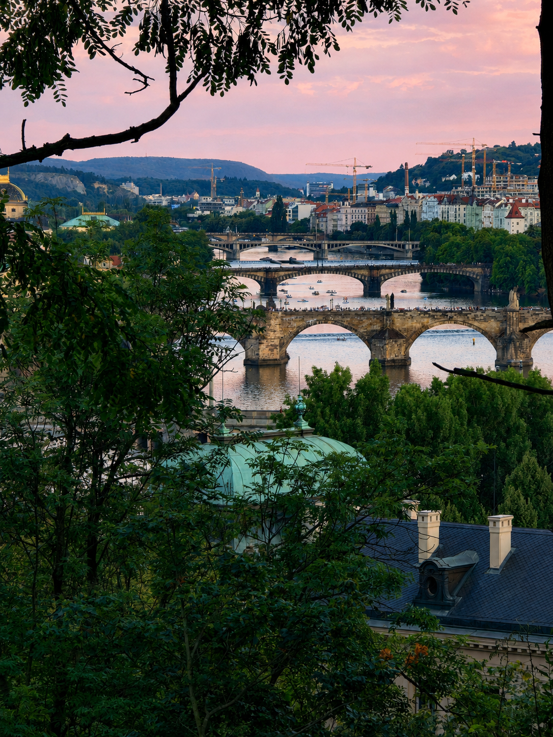 Prague bridges span the Vltava River beneath a pink evening sky, framed by trees and rooftops.