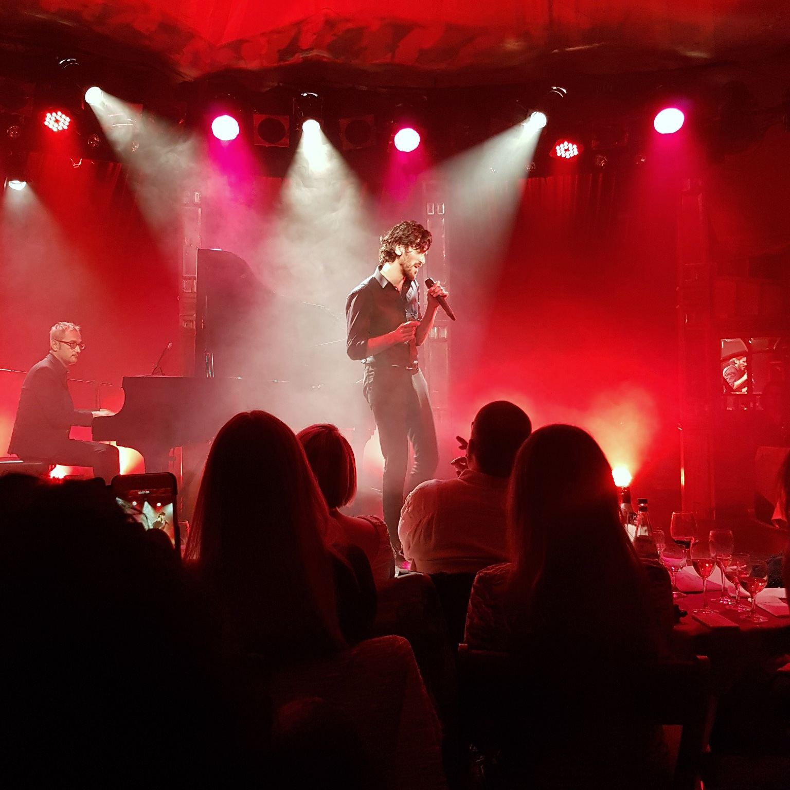 Singer Vladimir Korneev performs under red lights as a pianist plays, watched by seated audience members.