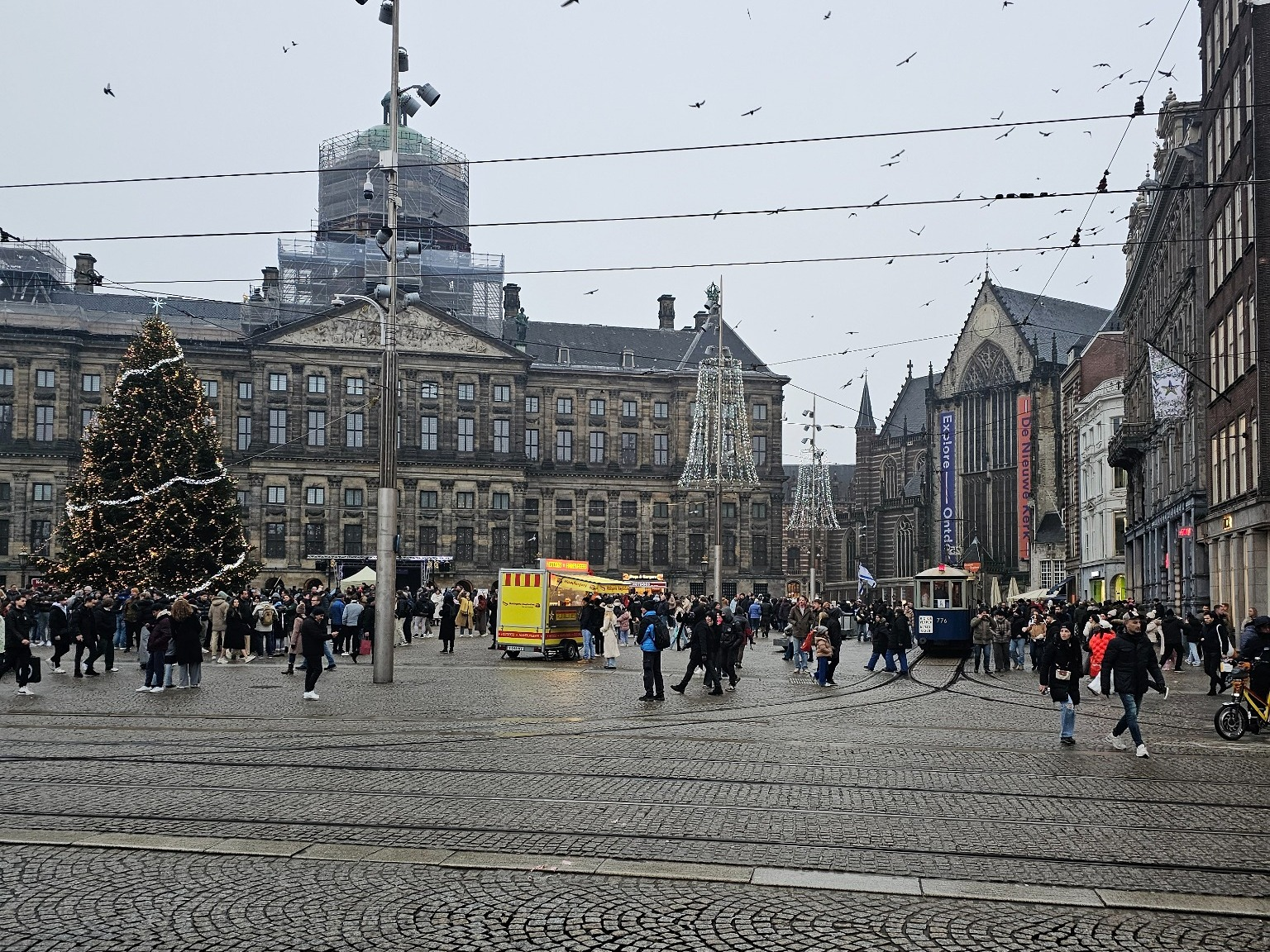 Dam Square in Amsterdam, with tram tracks, a Christmas tree, the Royal Palace, and crowds near the Nieuwe Kerk.