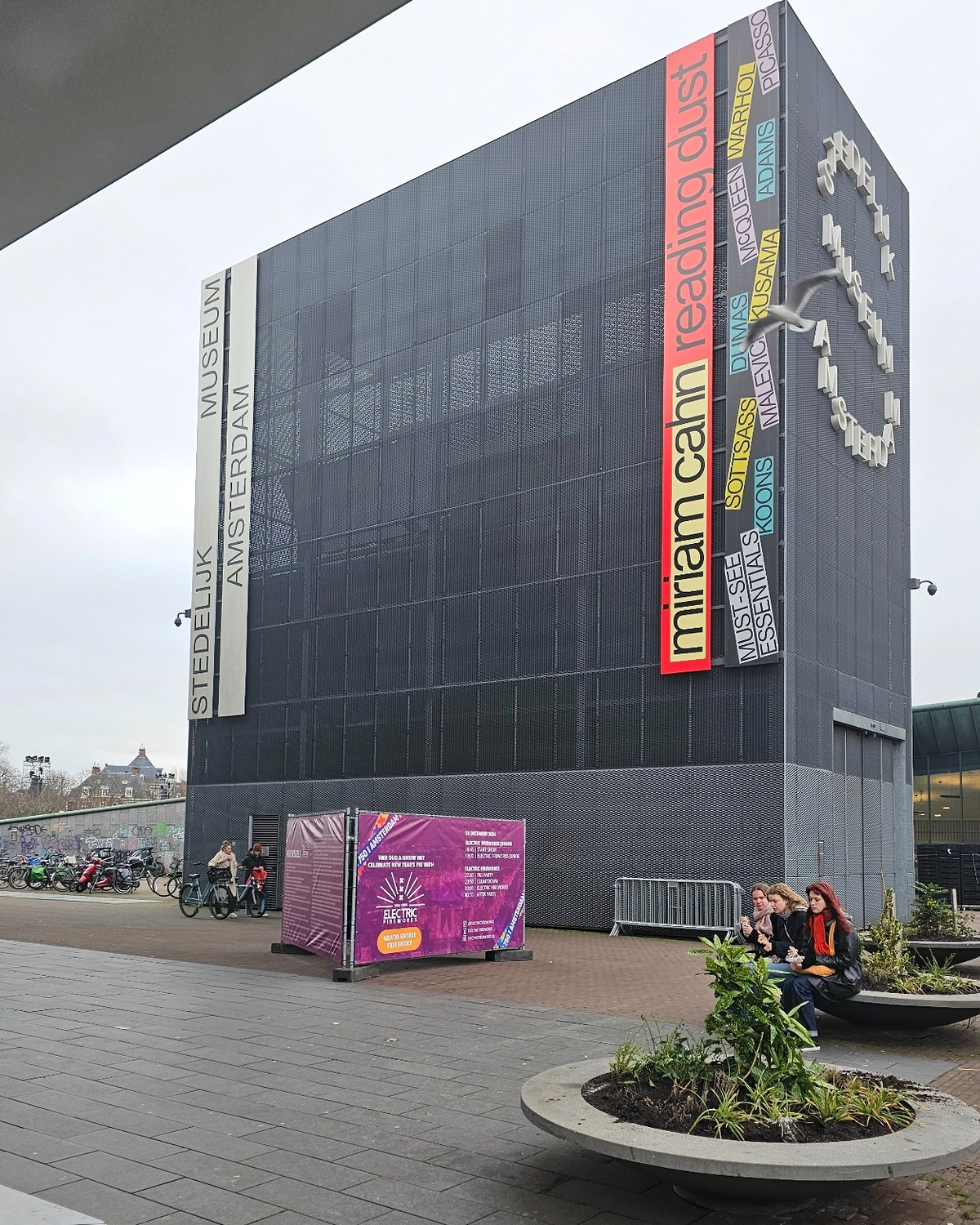 The Stedelijk Museum Amsterdam, wrapped in black mesh, displays colorful vertical banners and a purple sign outside.