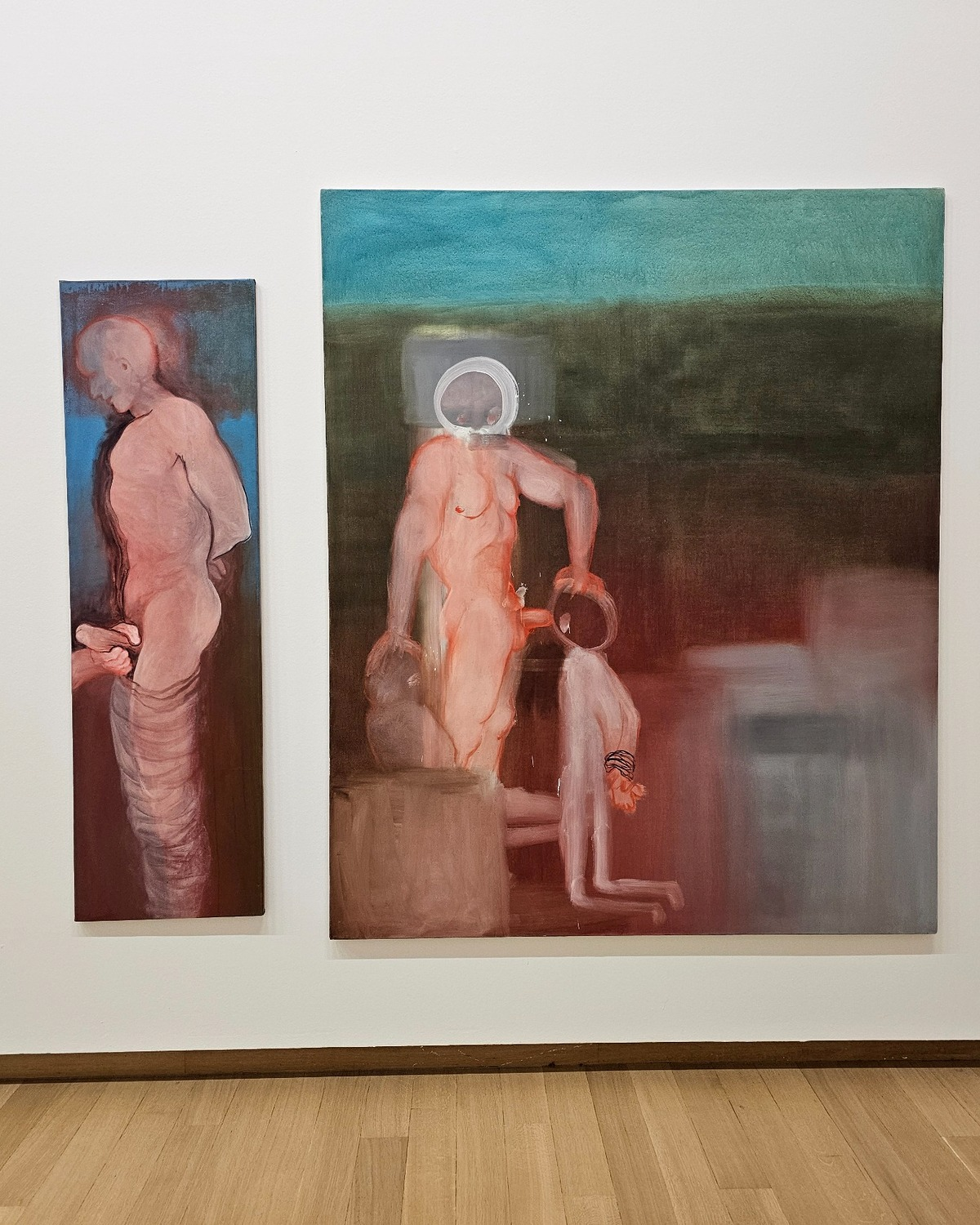 Two figurative paintings of human figures hang on a white gallery wall above a wooden floor.