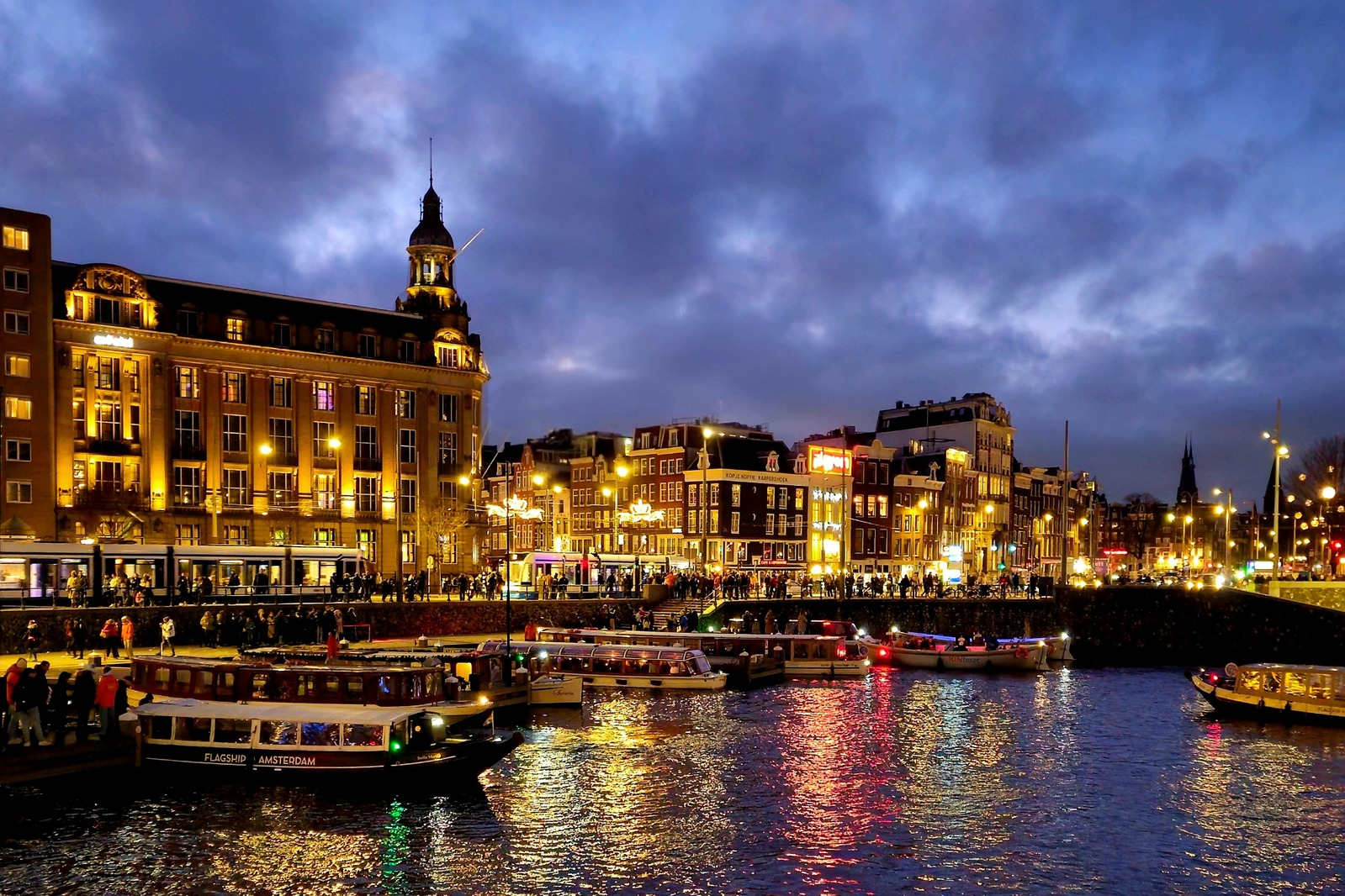 Illuminated Amsterdam canal with boats and canal-side buildings beneath a cloudy evening sky.