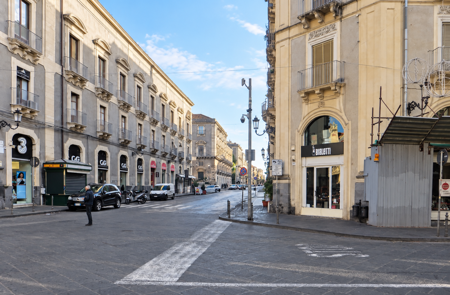 Catania street with pale Baroque buildings, shops, parked cars and dark stone paving.