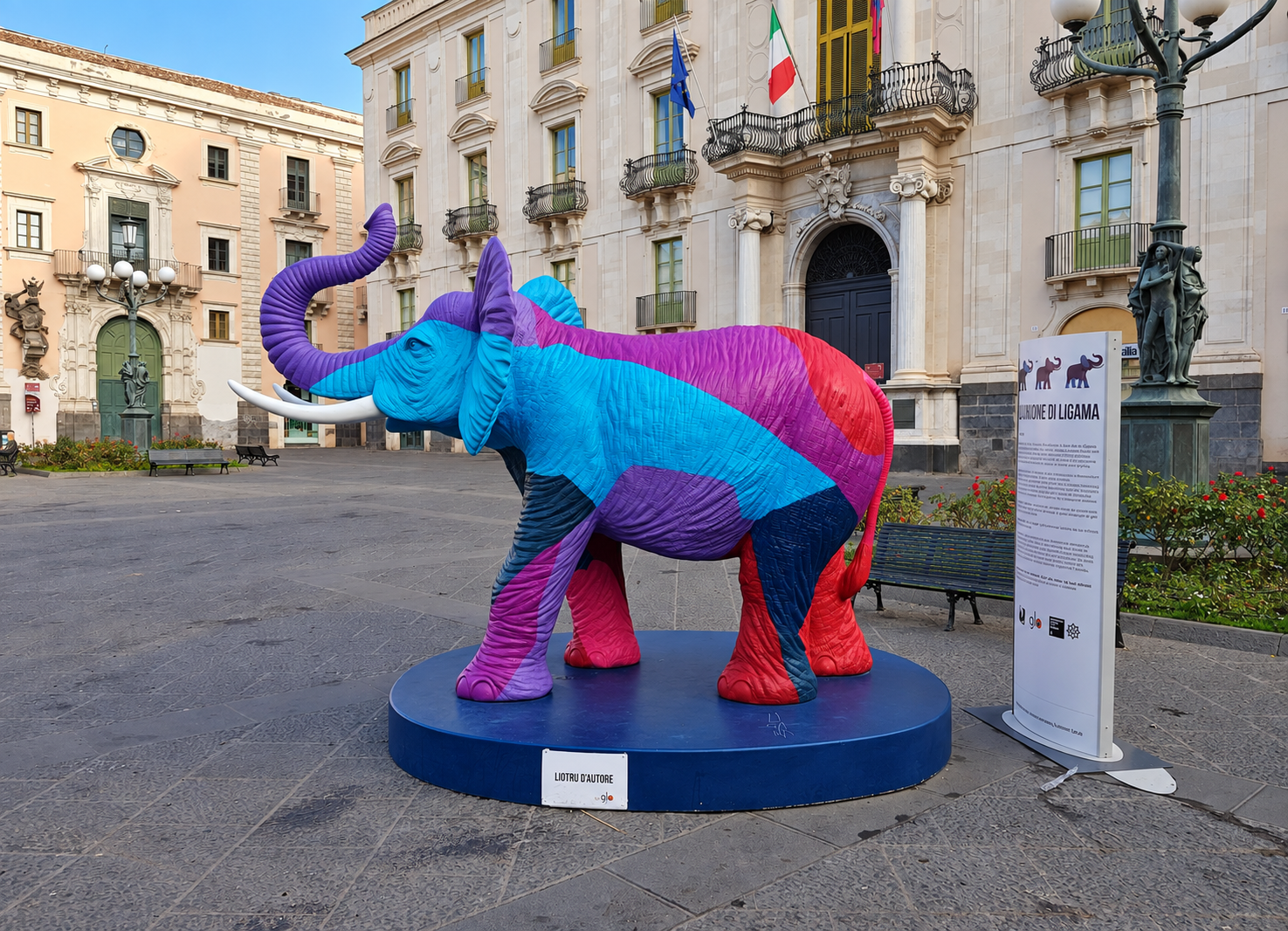 Colourful elephant sculpture in Piazza Università, Catania, with historic buildings and a sign behind it.