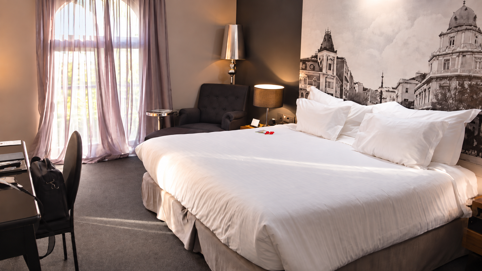 Hotel room with a large white bed, arched window, dark armchair, lamps and a black-and-white city mural.