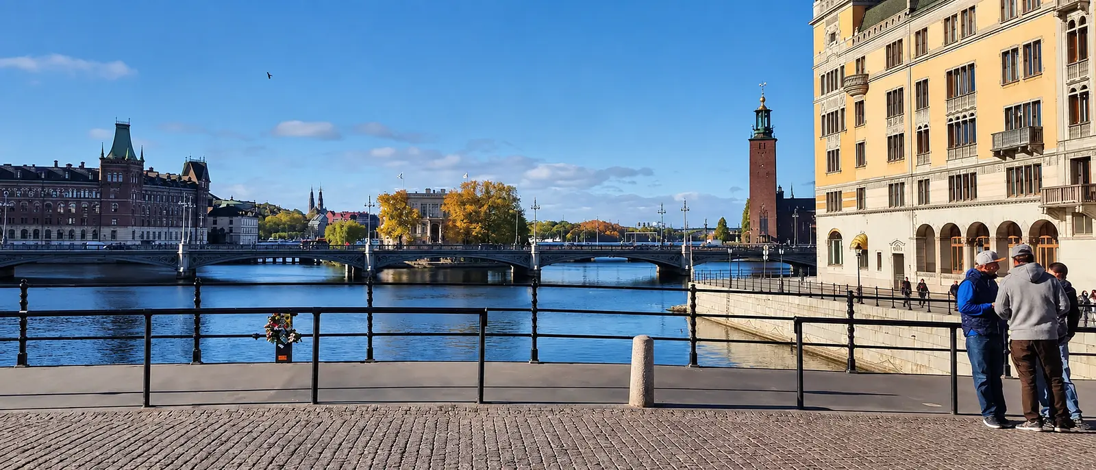 Stockholm waterfront with bridges, autumn trees, a tower, and two people beside the railing.