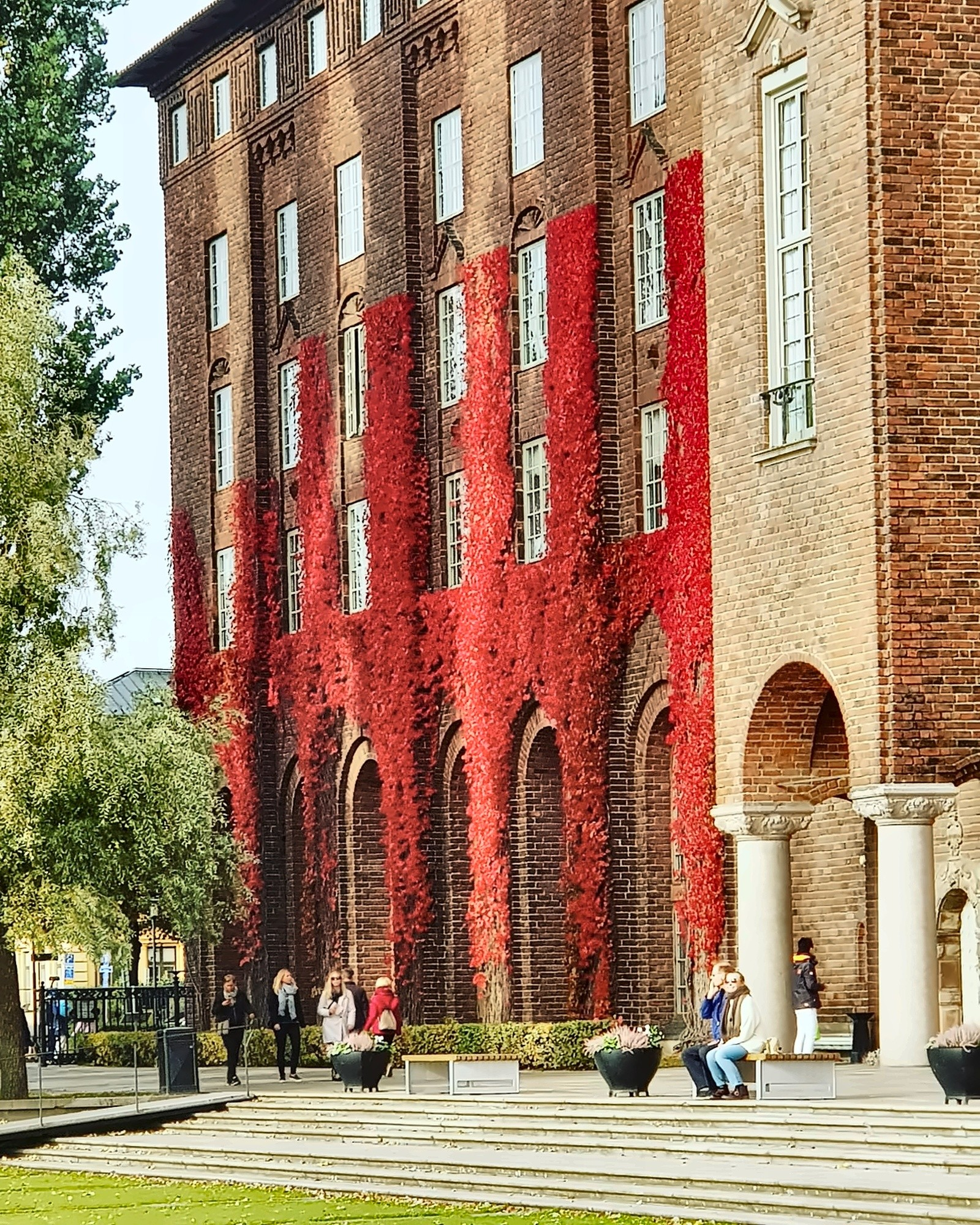 Red ivy covers the brick façade of Stockholm City Hall, with people on its front steps.