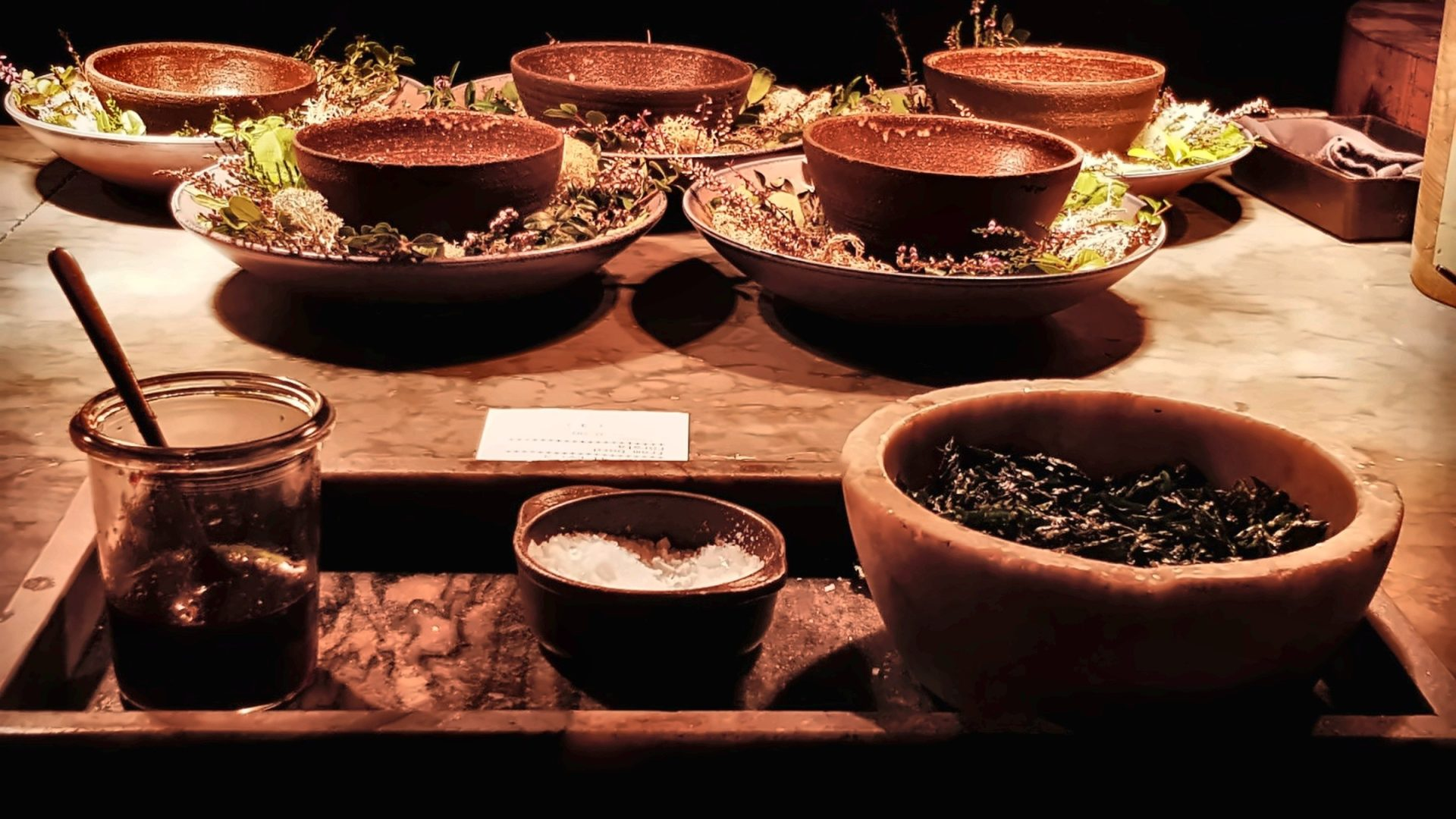 Bowls of reddish food and greens sit on a marble counter with a jar and spoon.