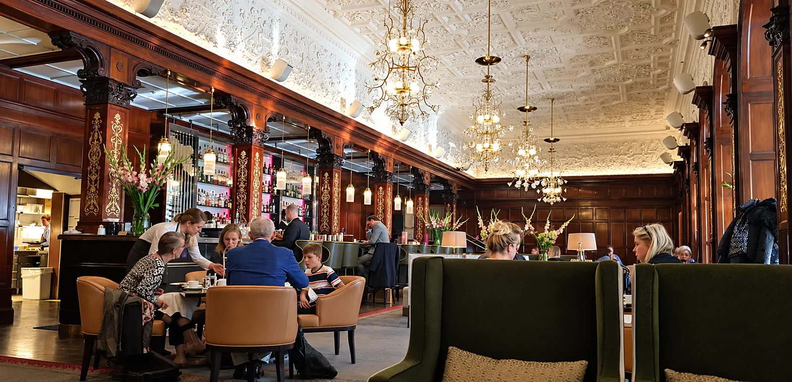 Diners sit in a wood-panelled Grand Hôtel salon beneath ornate chandeliers and a patterned ceiling.