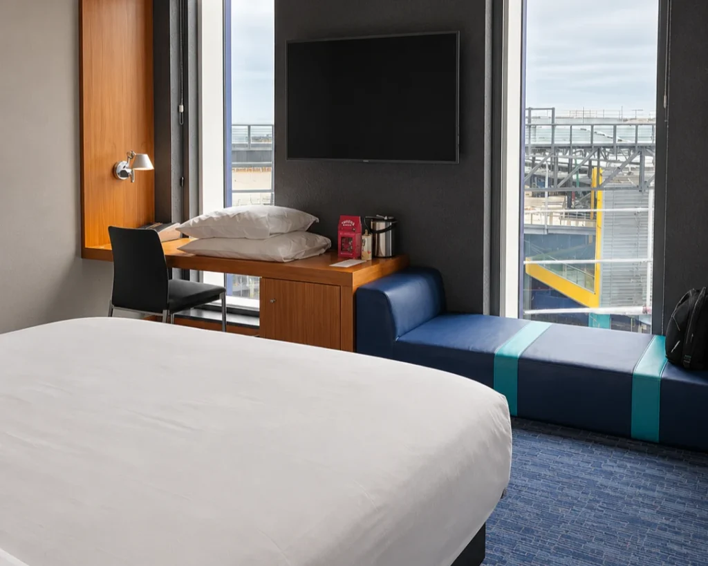 Hotel room with a bed, desk, television, and blue window seat overlooking an industrial structure.