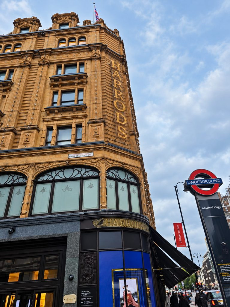 Harrods department store exterior beside a London Underground sign on a busy street.