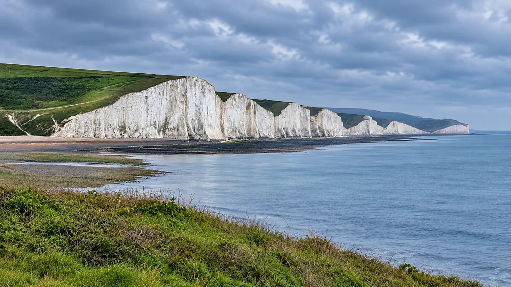White chalk cliffs of the Seven Sisters above the English Channel in East Sussex under a cloudy sky.