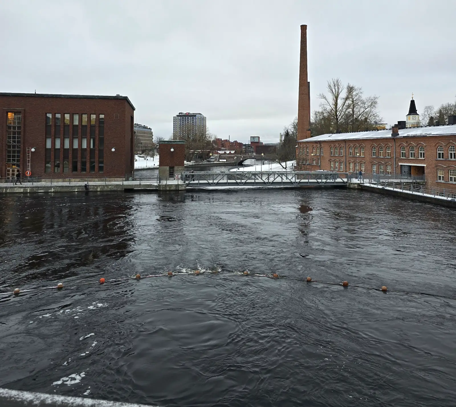 Tammerkoski rapids flow between snow-covered brick buildings and a footbridge in Tampere.