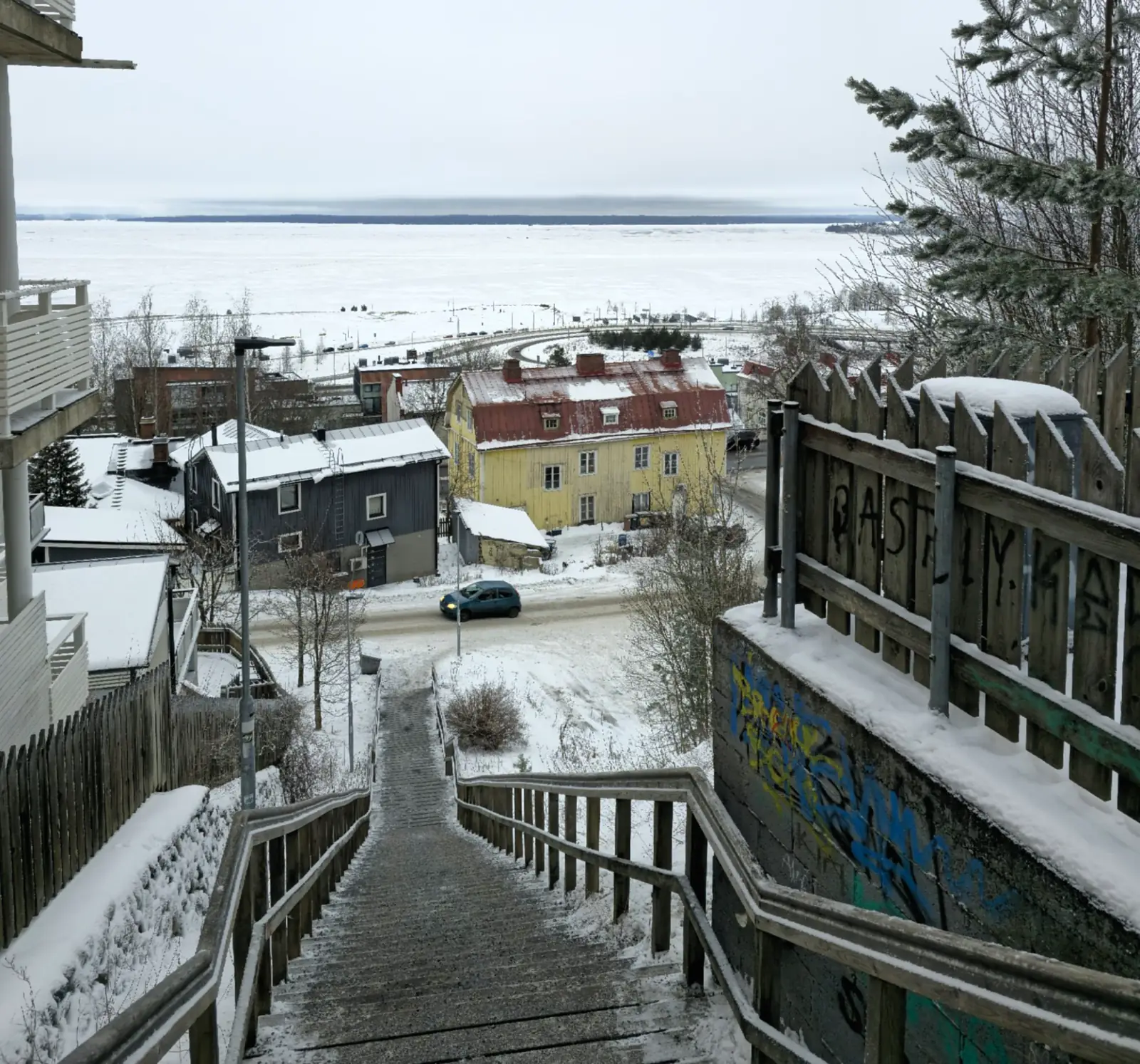 Snow-covered wooden stairs descend through Pispala, with homes and a frozen lake in the background.