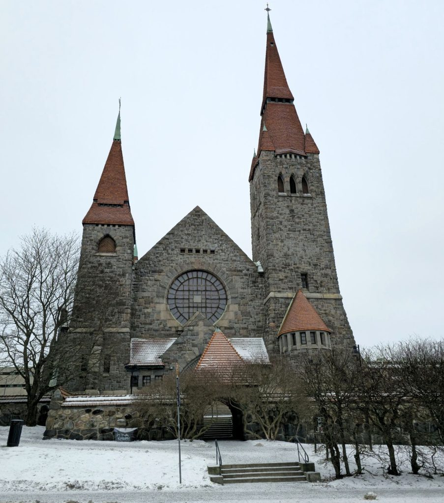 Tampere Cathedral stands in a snowy park, with gray stone walls and two red-roofed towers.