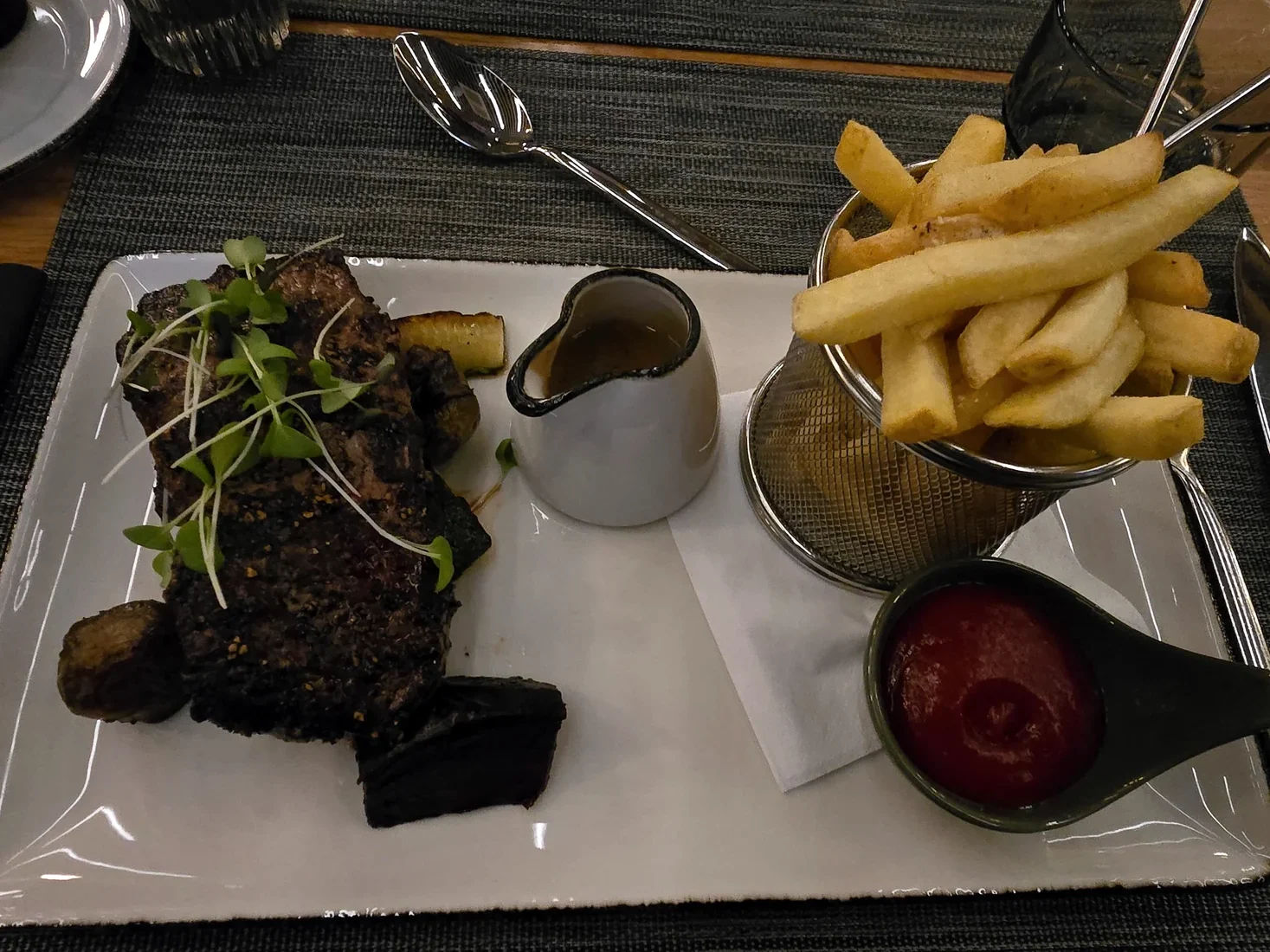 Grilled steak with microgreens, fries, gravy, and ketchup on a rectangular plate.