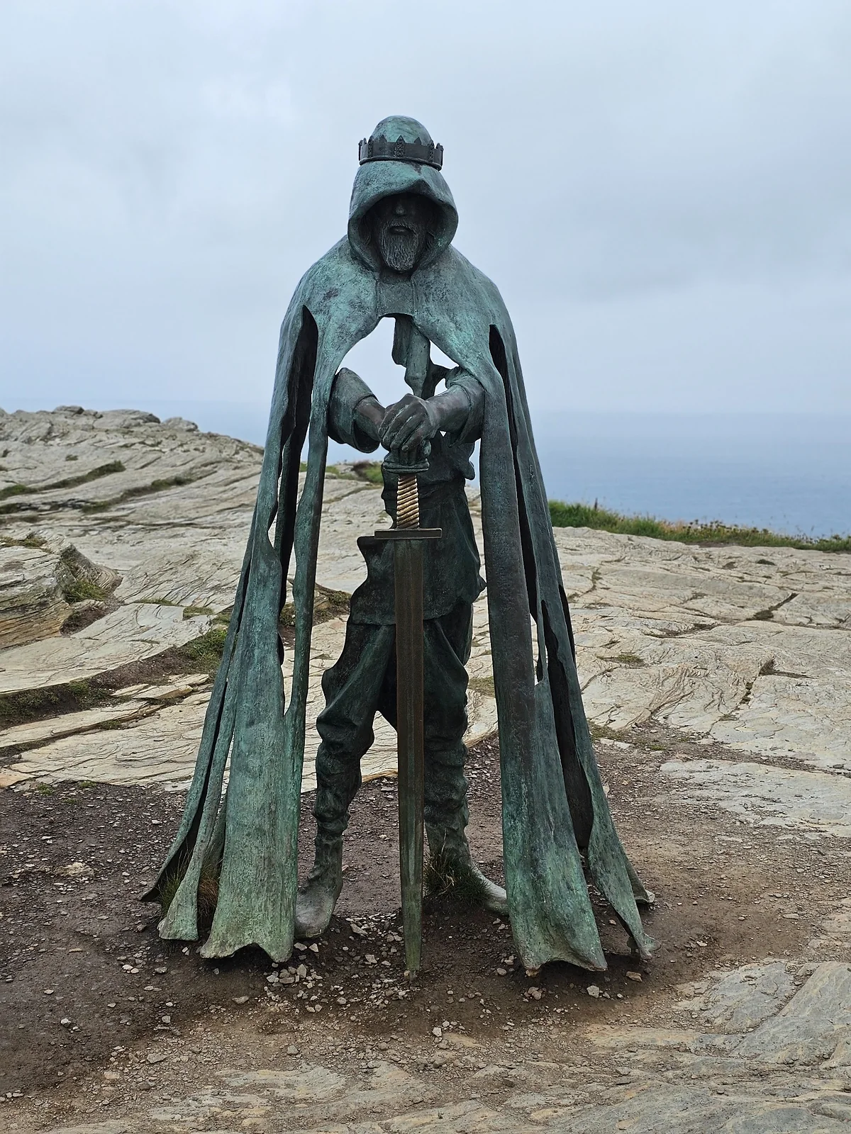 Bronze King Arthur statue overlooking the Atlantic at Tintagel, with rocky ground and a cloudy sky.