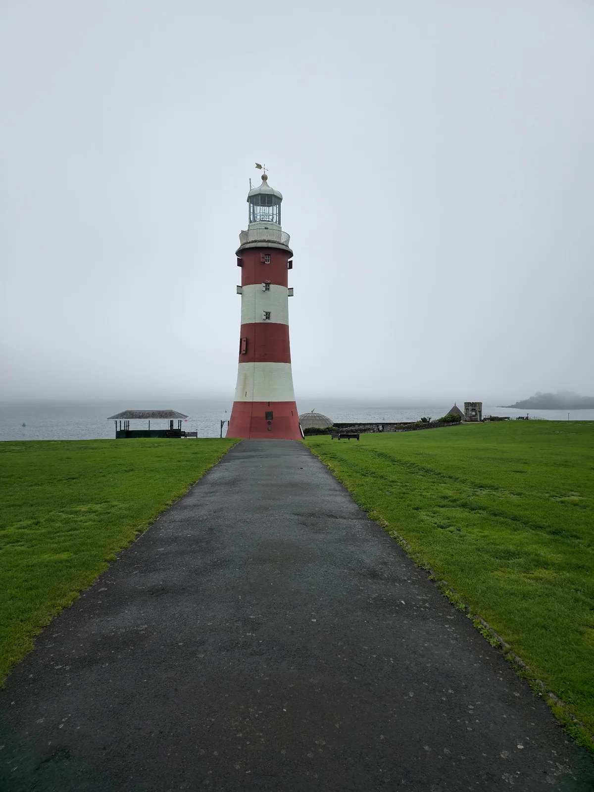 Smeaton’s Tower lighthouse stands at the end of a paved path on Plymouth Hoe beside the misty sea.