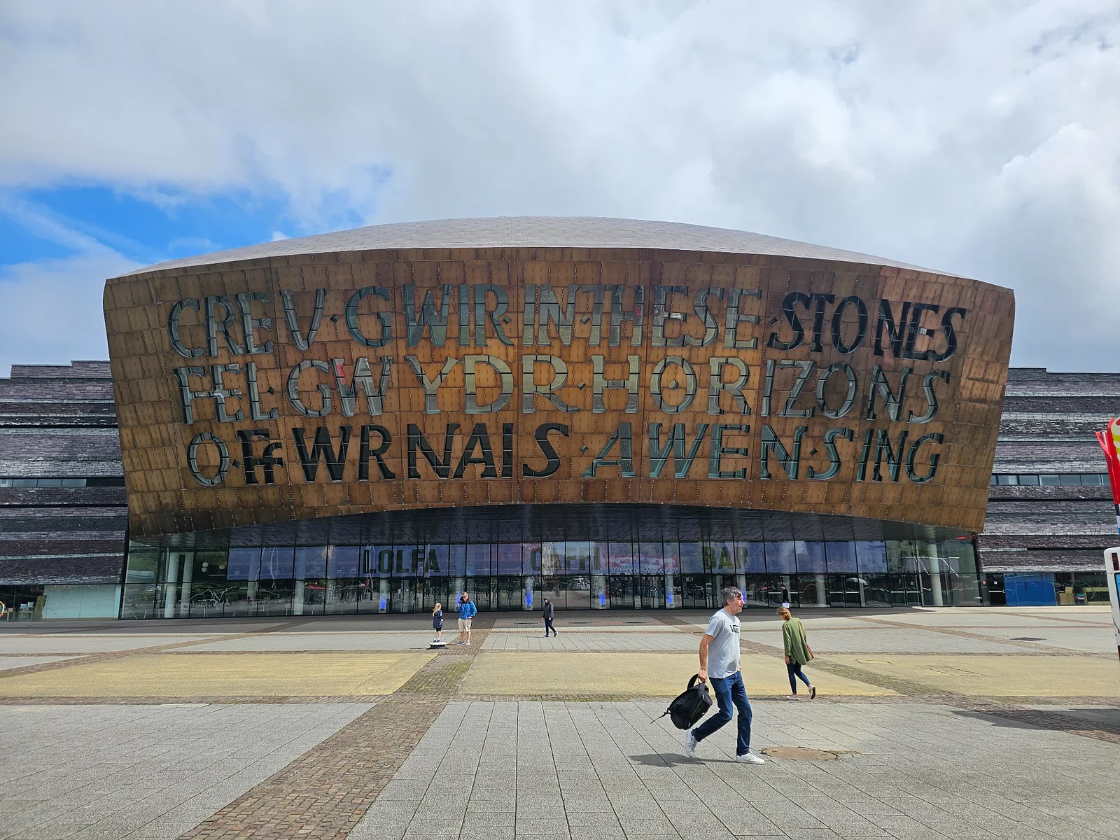Wales Millennium Centre in Cardiff Bay, with a bilingual inscription and people crossing the plaza.