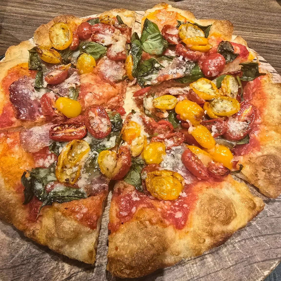 Pizza topped with cherry tomatoes, basil, cured meat, and grated cheese on a wooden table.