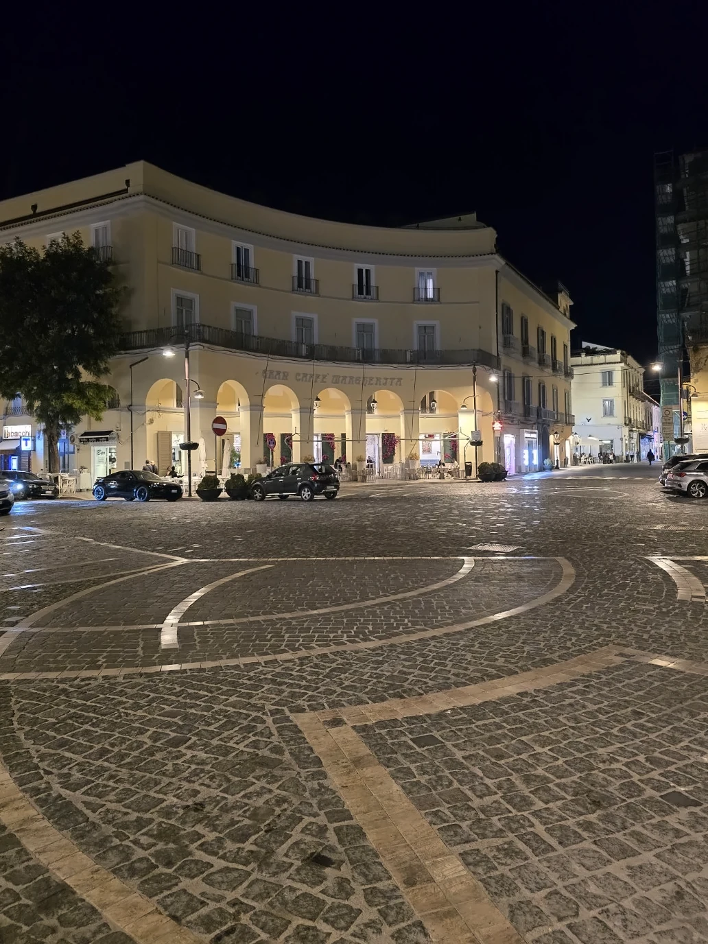 Piazza Dante at night, with an arcaded corner building, parked cars, and patterned cobblestones.