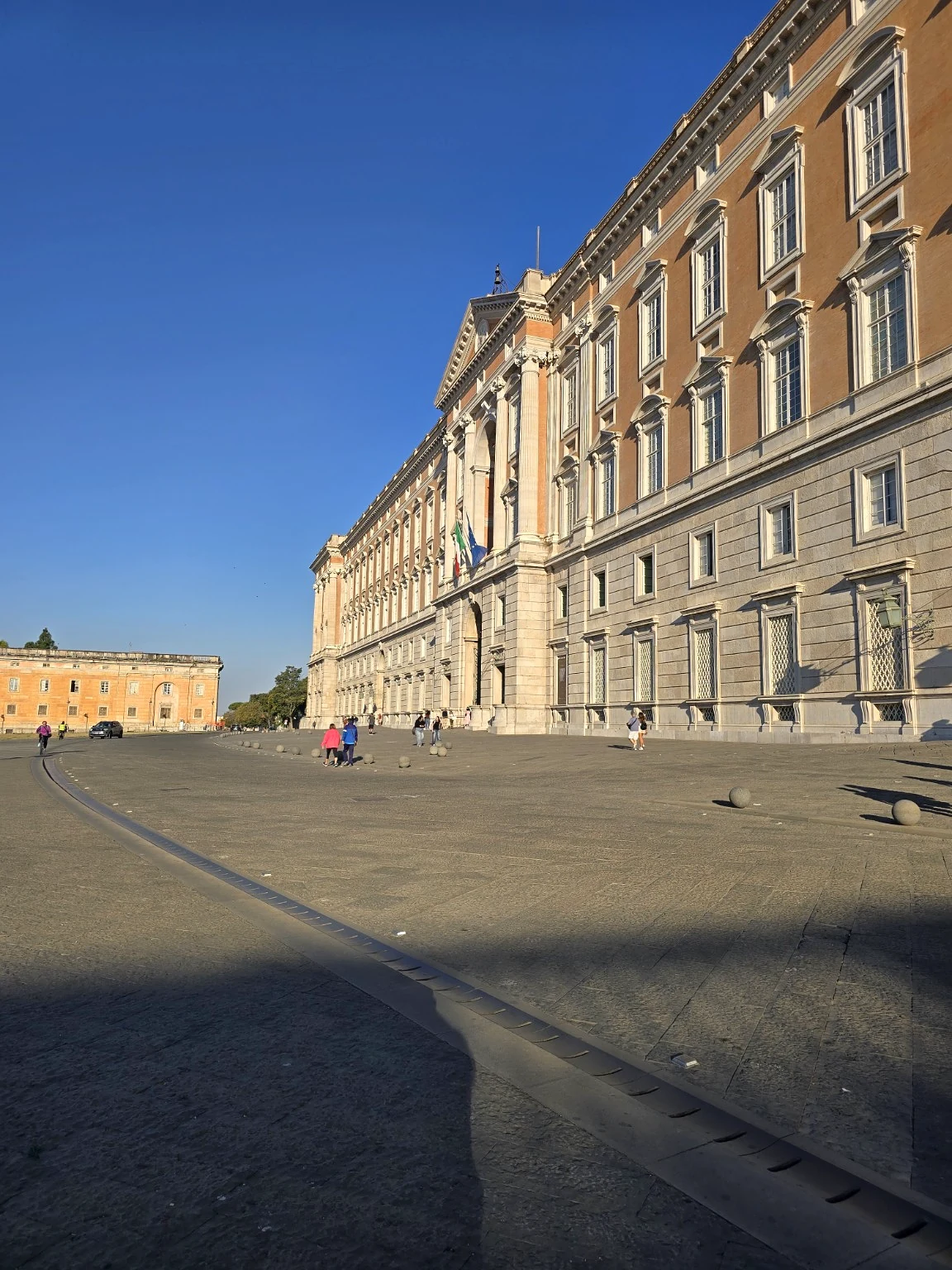 The Reggia di Caserta’s long stone facade borders a broad gravel courtyard with scattered visitors.