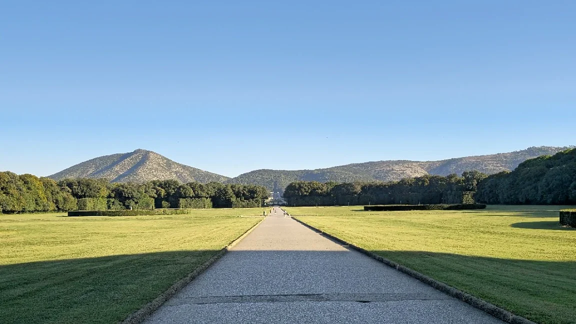 Long gravel avenue through the gardens of the Reggia di Caserta, with lawns, trees, and mountains beyond.