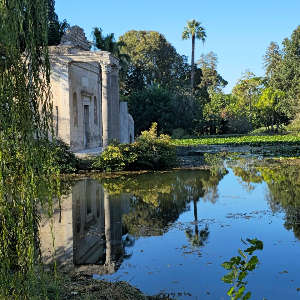 A stone garden pavilion beside a pond in the Reggia di Caserta gardens, reflected in the water.