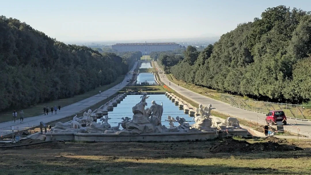 A fountain with marble statues sits in the Reggia di Caserta gardens, aligned with pools and the distant palace.