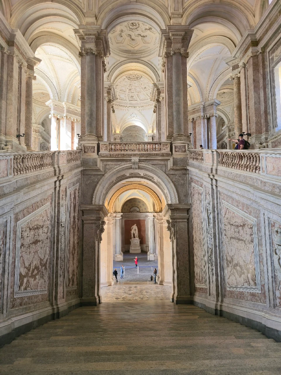 Ornate marble staircase inside the Reggia di Caserta, with arched ceilings and visitors below.