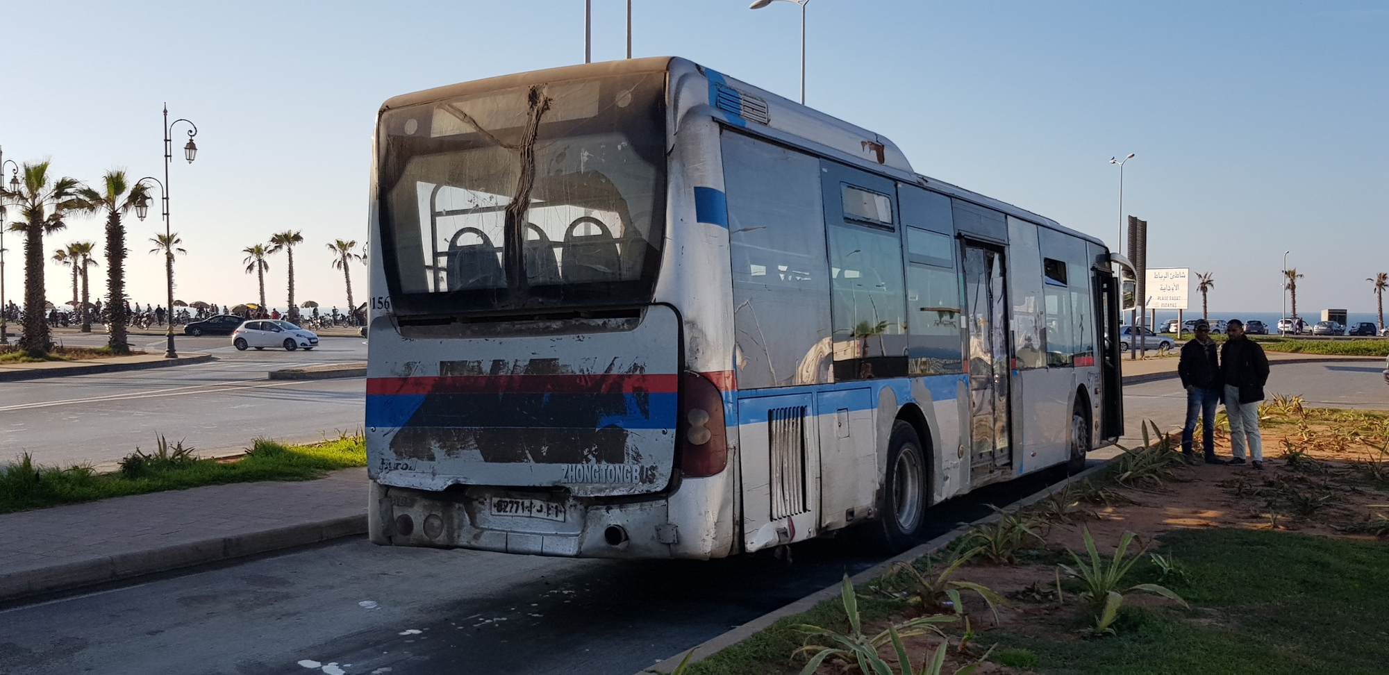 A weathered blue-and-white bus on a palm-lined road in Rabat, with two people standing beside it.