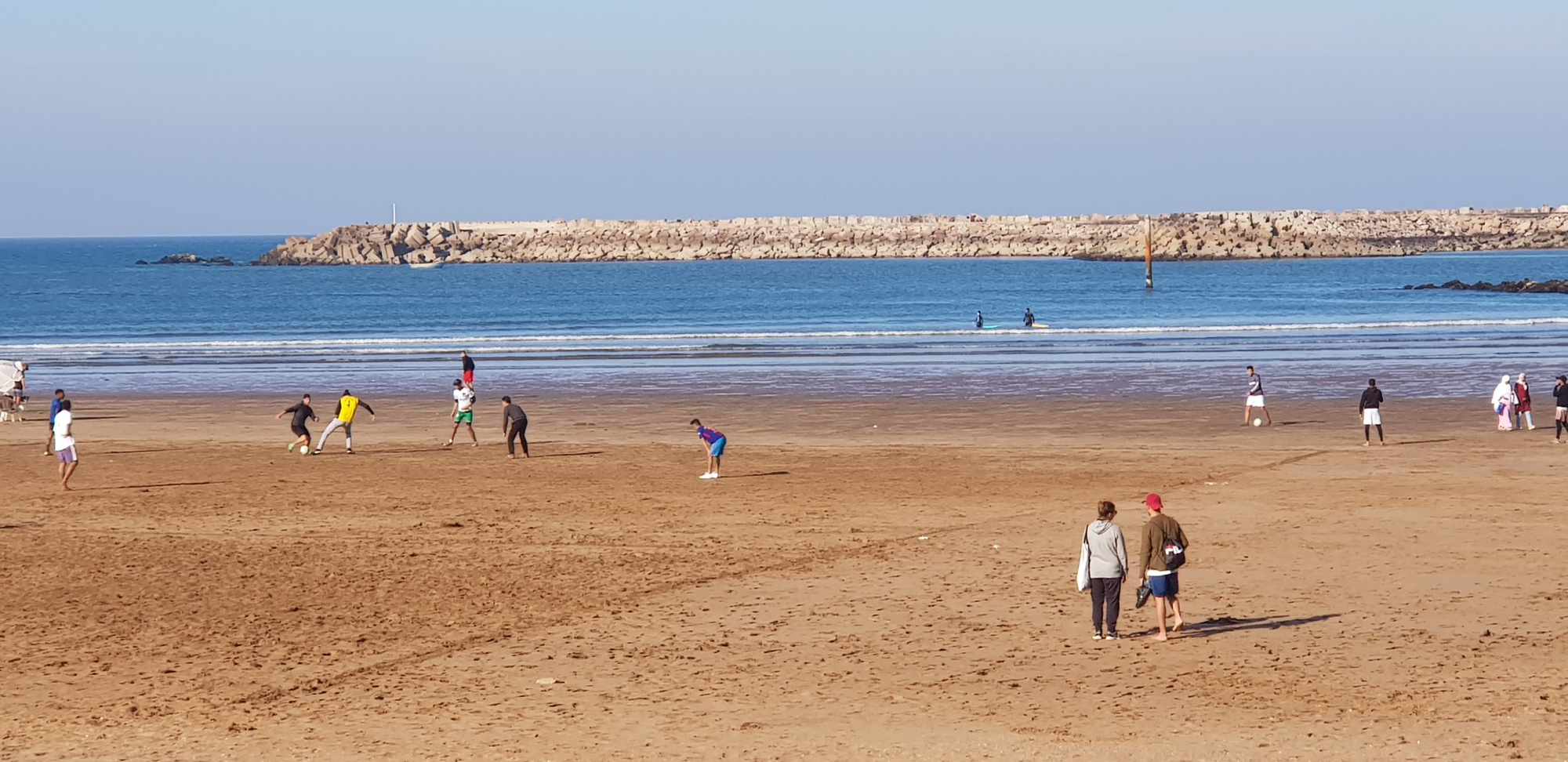 People walk and play on a sandy beach beside Rabat’s Atlantic waters and a rocky breakwater.