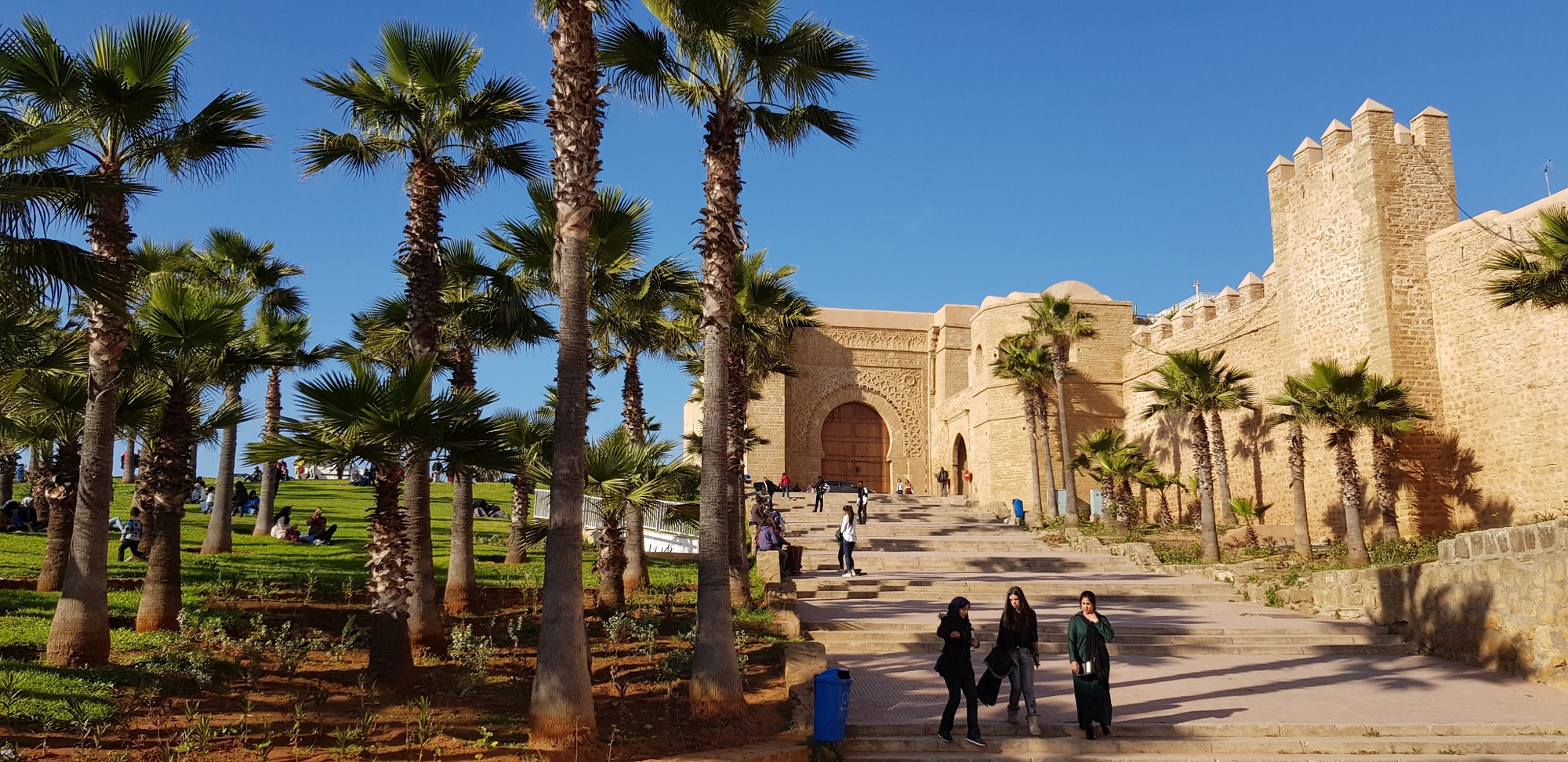 Palm-lined path leading to a stone fortress gate in Rabat, with several people walking nearby.